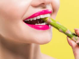 cheerful woman holding green and tasty asparagus in mouth isolated on yellow