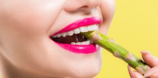 cheerful woman holding green and tasty asparagus in mouth isolated on yellow