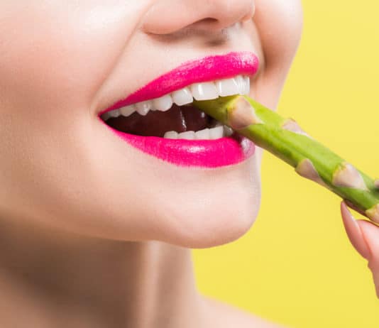 cheerful woman holding green and tasty asparagus in mouth isolated on yellow