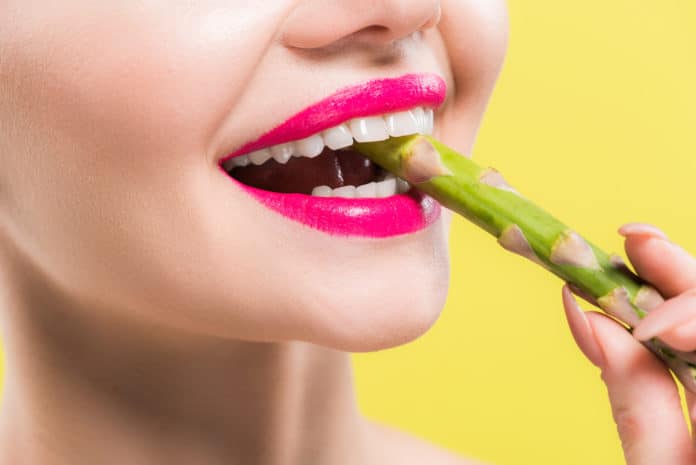 cheerful woman holding green and tasty asparagus in mouth isolated on yellow