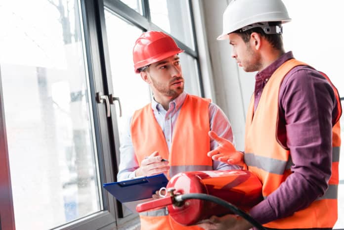 fireman in helmet looking at coworker while talking and holding red extinguisher