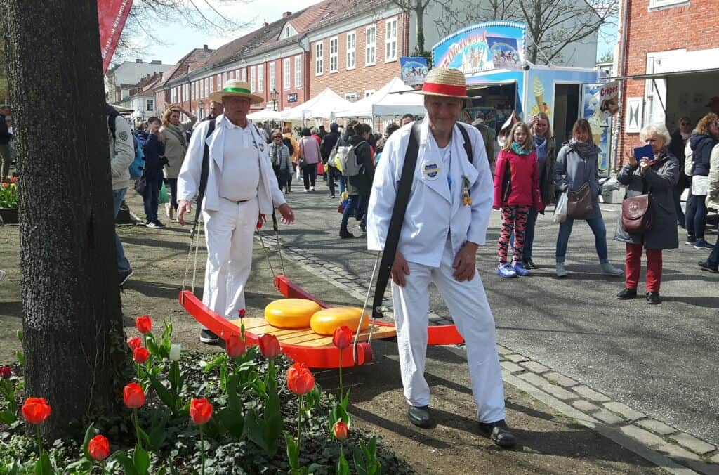 Zwei Männer in weißen Anzügen und Hüten beim Stadtfest mit Blumen und Besucher im Hintergrund.
