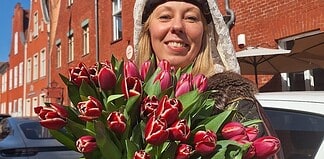 Blumenstrauß mit roten Tulpen, gehalten von einer lachenden Frau vor roten Backsteinhäusern.