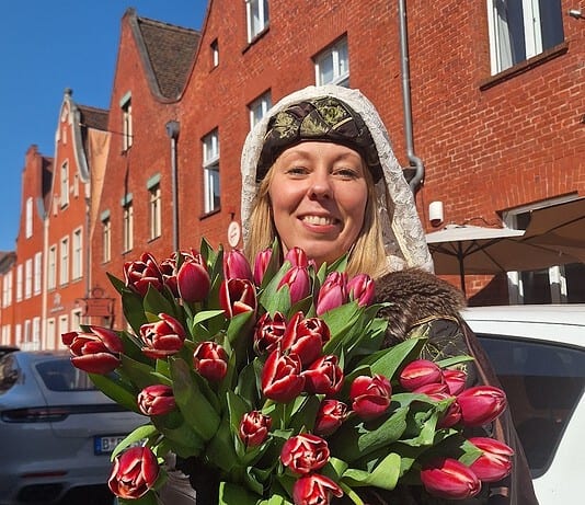 Blumenstrauß mit roten Tulpen, gehalten von einer lachenden Frau vor roten Backsteinhäusern.