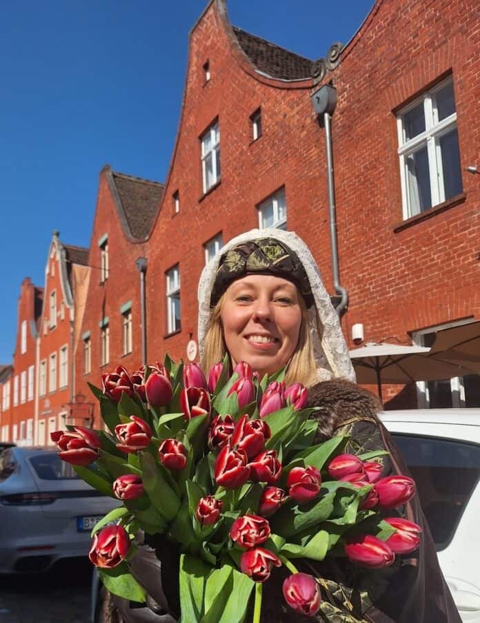 Blumenstrauß mit roten Tulpen, gehalten von einer lachenden Frau vor roten Backsteinhäusern.